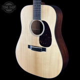 A close-up studio shot of an acoustic guitar against a solid black background, featuring a natural wood grain top, dark brown sides, and a black pickguard. A Tone Shop watermark is visible in the upper left corner.