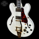 A white electric guitar with a glossy finish, featuring a red tortoiseshell pickguard, gold hardware, and black controls, shown against a solid black background.