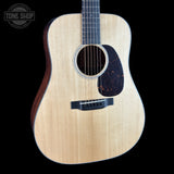A close-up studio shot of an acoustic guitar body against a solid black background, featuring a natural wood grain top, a dark pickguard, and a black bridge. A Tone Shop logo is visible in the top-left corner.