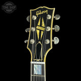 Close-up of a Gibson guitar headstock with six tuning pegs, featuring the gold Gibson logo and a gold geometric emblem on a black background.