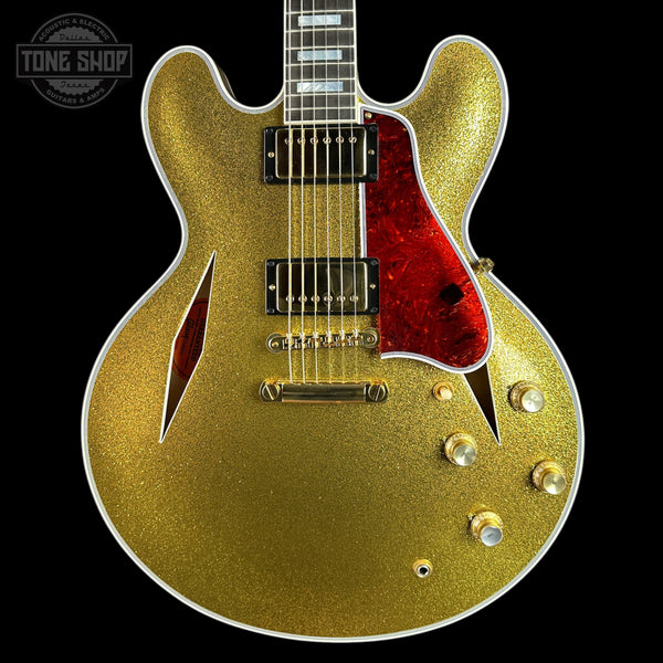 A gold glitter electric guitar with a red pickguard and black pickups, shown against a solid black background with a Tone Shop watermark in the top left corner.