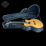 An acoustic guitar with a natural wood finish resting inside an open black hard case on a black background. A Tone Shop watermark is visible in the top-left corner.