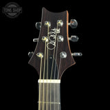 Close-up of the headstock of a dark wood acoustic guitar against a black background, featuring a white script signature and six tuning pegs.