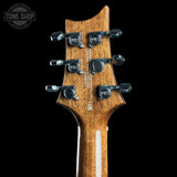 Close-up of the headstock of an electric guitar, showing the natural wood grain, six black tuning pegs with silver screws, and the PRS logo on the tuning machines. The background is solid black.