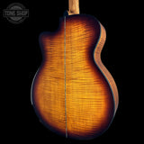 Close-up of the back of an acoustic guitar showing the rich, warm sunburst wood grain finish on a solid black background. A Tone Shop watermark is visible in the top-left corner.