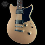 A close-up of an electric guitar body in a metallic champagne gold finish with a black pickguard, black humbucker pickups, and chrome hardware, shown against a solid black background.