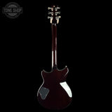 A dark reddish-brown electric guitar shown from the back against a solid black background, with a white circular badge on the lower back and a Tone Shop watermark in the top left corner.