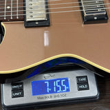 Close-up of a guitar body being weighed on a digital scale, with the scale display reading 7.155 lbs and the guitar showing a metallic brown finish and black pickups.