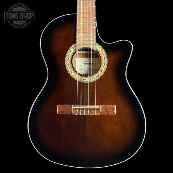 An acoustic guitar with a dark brown sunburst finish, featuring a cutaway body, white binding, and a visible Ibanez logo on the soundboard, set against a solid black background.
