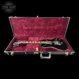 A black electric guitar displayed inside an open hard case with a deep magenta velvet interior, photographed against a solid black background. A Tone Shop watermark is visible in the top-left corner.