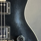 Close-up of a black electric guitar body showing the glossy finish, white binding, part of a pickup, strings, and a control knob against a neutral gray background.