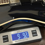 A close-up of a glossy black electric guitar body resting on a digital scale displaying 9.2 oz.