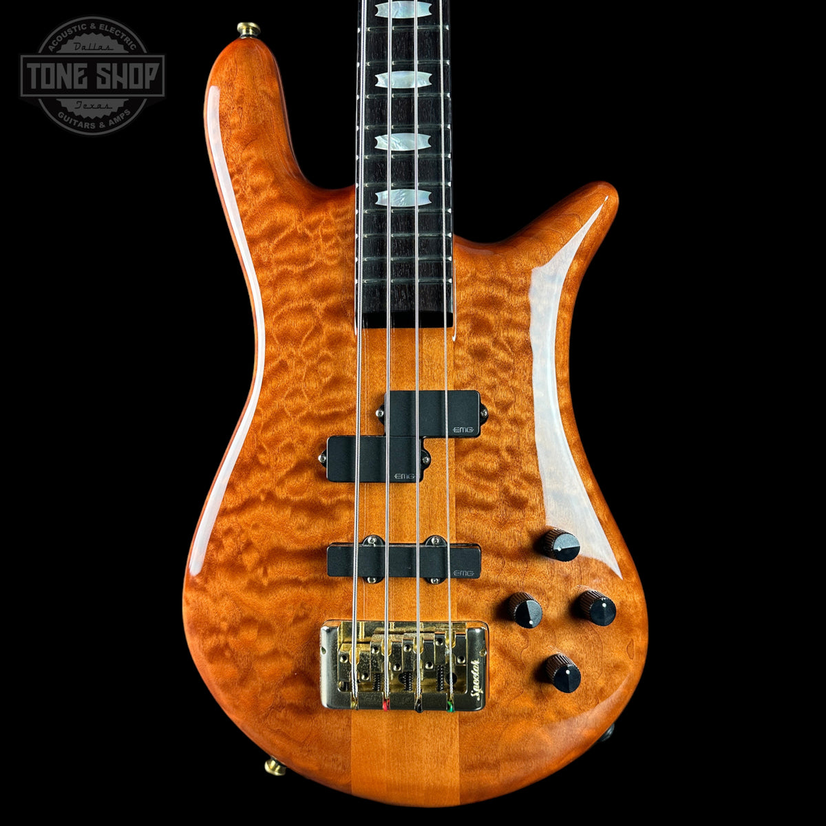 A close-up of a four-string bass guitar with a vibrant orange flamed maple wood finish, black hardware, and a black neck, shown against a solid black background with a Tone Shop watermark in the top left corner.