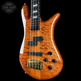 A close-up of a four-string bass guitar with a glossy orange-brown flamed maple top, black hardware, and a black neck, shown against a solid black background. A Tone Shop watermark is visible in the top-left corner.