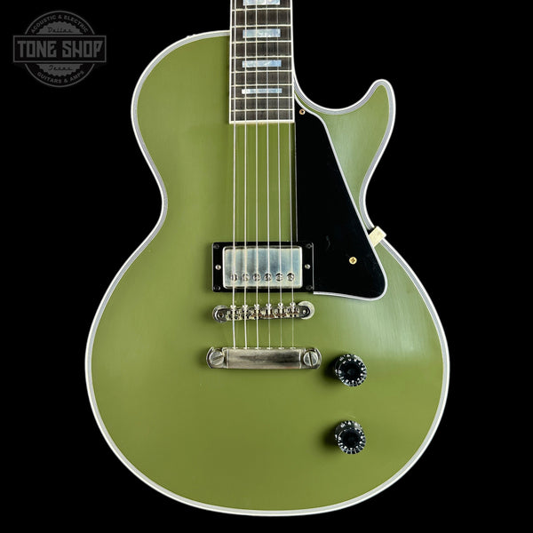 An electric guitar with an olive green body, black pickguard, chrome hardware, and a black-and-white striped fretboard, shown against a solid black background with a Tone Shop watermark in the top left corner.