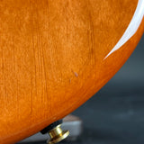 Close-up detail of a curved wooden body, likely an electric guitar, showing a warm orange-brown wood finish, a white edge stripe, and a brass hardware component against a dark blurred background.