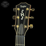 Close-up of a Taylor acoustic guitar headstock with six gold tuning pegs, gold strings, and a white scroll logo on a black background.