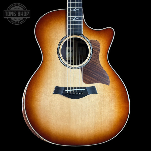 A Taylor acoustic guitar with a natural sunburst finish, shown from the front against a solid black background. The brand logo is visible on the headstock (partially cropped) and the Tone Shop watermark is in the top left corner.