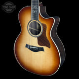 A Taylor acoustic-electric guitar with a natural sunburst finish, dark wood sides, and a cutaway body, shown against a solid black background with a Tone Shop watermark in the top left corner.