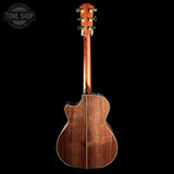 The back of a wooden acoustic-electric guitar, showing a rich reddish-brown wood grain finish and gold tuning pegs, photographed against a solid black background with a Tone Shop watermark in the top-left corner.