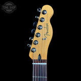 Close-up of the headstock of a Fender electric guitar, showing the maple wood headstock, six tuning pegs, and the Fender logo, set against a solid black background.
