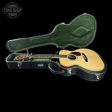An acoustic guitar with a natural wood top and dark neck, displayed inside an open black hard case with green felt lining, set against a solid black background.