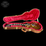 A gold-finish electric guitar inside an open brown hard case with bright red plush lining, photographed on a black background. A Tone Shop watermark appears in the upper left corner.