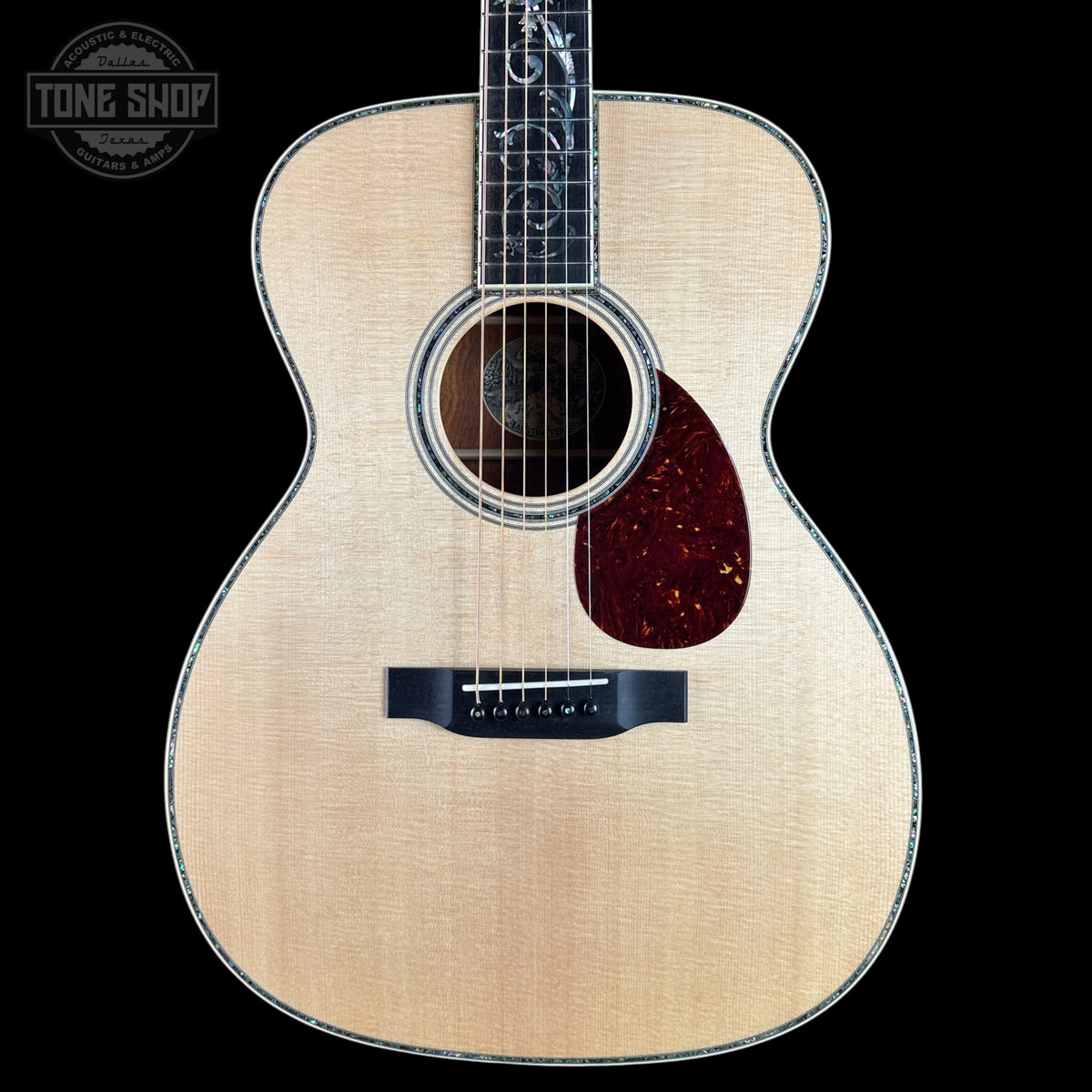 A front-facing acoustic guitar with a natural wood finish and dark pickguard, shown against a solid black background with a Tone Shop logo in the top-left corner.