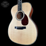 A natural wood acoustic guitar with a dark reddish-brown pickguard and black bridge, shown against a solid black background. A Tone Shop logo is visible in the top-left corner.