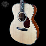 An acoustic guitar with a natural wood top, dark brown sides, and decorative inlays on the fretboard, shown against a solid black background with a Tone Shop watermark in the top left corner.