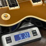 Close-up of a gold electric guitar body resting on a digital scale displaying 9.22 lbs, with the guitar's cream-colored binding and pickups visible.