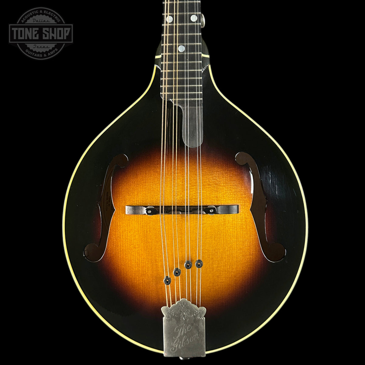 A close-up studio shot of a mandolin against a solid black background, featuring a classic sunburst finish that transitions from black at the edges to a warm amber-orange center. The mandolin has a silver metal tailpiece and bridge, with a Tone Shop logo watermark in the upper left corner.