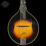 A close-up studio shot of a mandolin against a solid black background, featuring a classic sunburst finish that transitions from black at the edges to a warm amber-orange center. The mandolin has a silver metal tailpiece and bridge, with a Tone Shop logo watermark in the upper left corner.