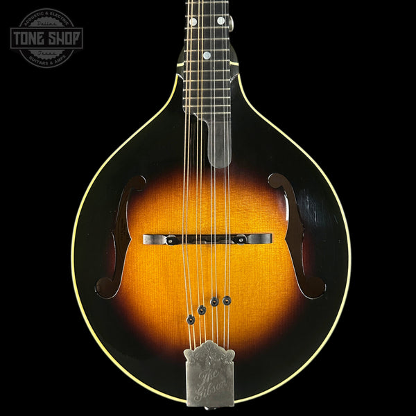 A close-up studio shot of a mandolin against a solid black background, featuring a classic sunburst finish that transitions from black at the edges to a warm amber-orange center. The mandolin has a silver metal tailpiece and bridge, with a Tone Shop logo watermark in the upper left corner.