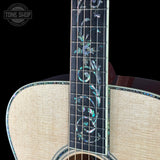 Close-up of an acoustic guitar showing the neck with intricate iridescent mother-of-pearl floral inlays on a black fretboard, alongside the light natural wood body and matching inlaid edges, set against a solid black background.