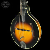 A close-up studio shot of a mandolin against a solid black background, featuring a classic sunburst finish that transitions from black to amber-orange, with a white binding edge. The brand 'The Gibson' is visible on the metal tailpiece, and a 'TONE SHOP' watermark is in the top-left corner.