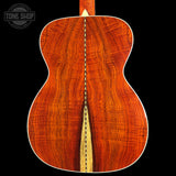 Close-up of the back of an acoustic guitar showing a rich, wavy wood grain pattern in warm reddish-brown tones, with a light natural wood center stripe and white binding, set against a solid black background.