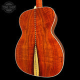 The back of a acoustic guitar made of rich, warm-toned wood with a natural sound hole and a white binding edge, shown against a solid black background with a Tone Shop watermark in the top left.