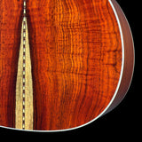 Close-up of an acoustic guitar body showing a rich flamed wood grain finish in warm reddish-brown tones, with a decorative inlay running vertically and a white edge binding against a black background.