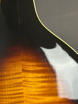Close-up of a glossy acoustic guitar body showing a dark top edge, a pale binding line, and a flamed sunburst wood finish shifting from black to amber and orange.