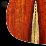 Close-up detail of an acoustic guitar's soundboard, showcasing a rich reddish-brown wood grain with a decorative inlay running vertically down the center. The tone is well-lit against a solid black background, with a Tone Shop watermark in the upper left corner.