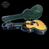 An acoustic guitar with a natural wood top and dark neck, displayed inside an open black hard case with green felt lining, set against a solid black background.