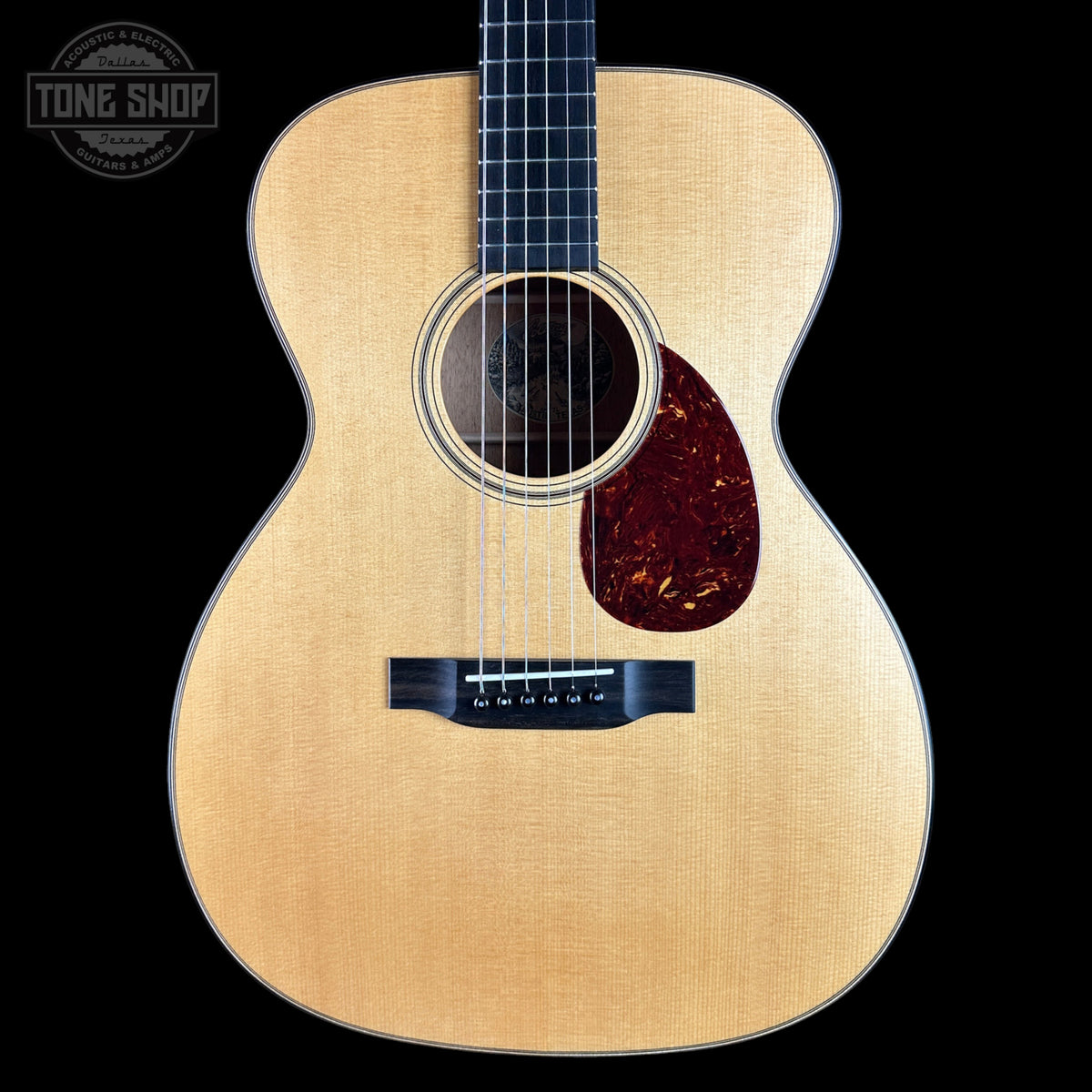 A close-up studio shot of an acoustic guitar body against a solid black background, featuring a natural wood finish, a dark reddish-brown pickguard, and a Tone Shop watermark in the upper left corner.