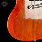 Close-up of the body of an electric guitar with a glossy reddish-brown sunburst finish, showing the wood grain and a portion of the chrome bridge against a black background.
