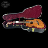An acoustic ukulele with a natural wood body and dark neck, displayed inside an open black hard case with a burgundy velvet lining, set against a solid black background.