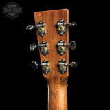 Close-up of a guitar headstock made of warm brown wood with six tuning pegs and decorative metal hardware, shown against a black background. A Tone Shop watermark is visible in the top left corner.