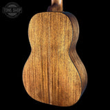 Close-up of the back of a wooden ukulele or guitar showing a rich grain pattern in warm brown and golden tones against a solid black background. A Tone Shop watermark is visible in the top left corner.