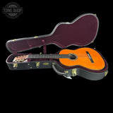 An acoustic guitar with a bright orange spruce top is displayed inside an open black hard case with a burgundy velvet lining. The Tone Shop logo is visible in the top-left corner.