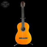 A classical guitar with a vibrant orange-yellow sunburst finish, displayed on a black background with a metal stand. A 'TONE SHOP' watermark is visible in the top-left corner.