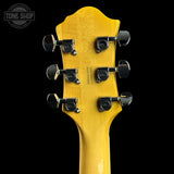 Close-up of the headstock of a guitar, showing six black tuning pegs and a bright yellow maple wood backplate against a black background. A Tone Shop watermark is visible in the top left corner.